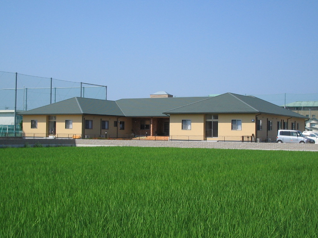 広大な田園に囲まれた平屋建ての介護施設の外観。明るい外壁と緑の屋根が特徴。